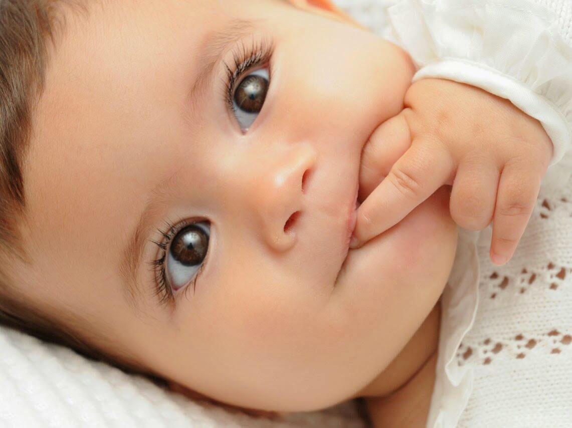 A baby with brown eyes putting their fingers in their mouth while lying down.