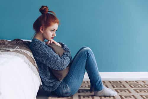 En teenagepige sidder på gulvet ved siden af sin seng og krammer en pude