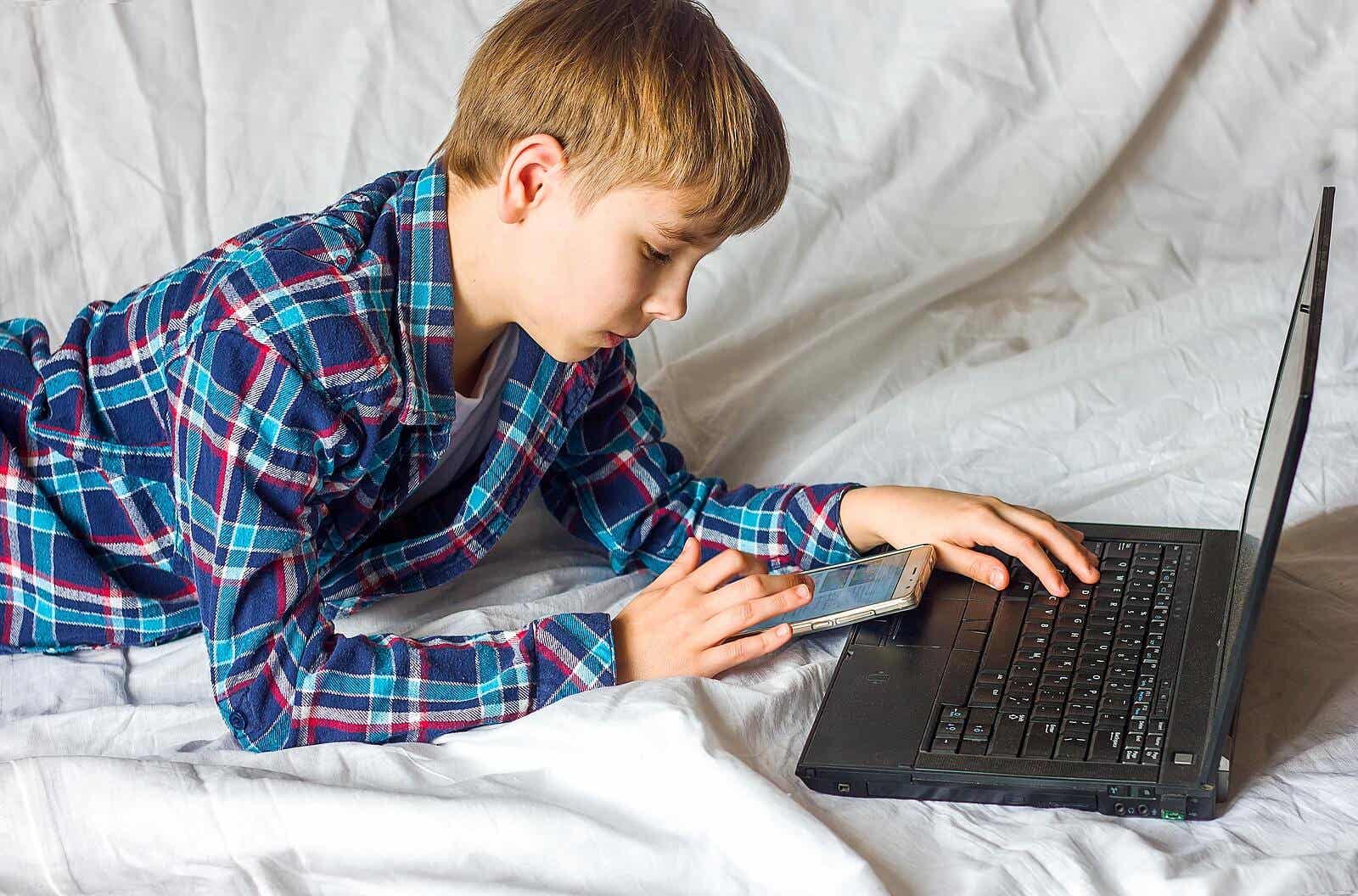 Een tiener die naar zijn mobiele telefoon kijkt terwijl hij op de computer werkt.