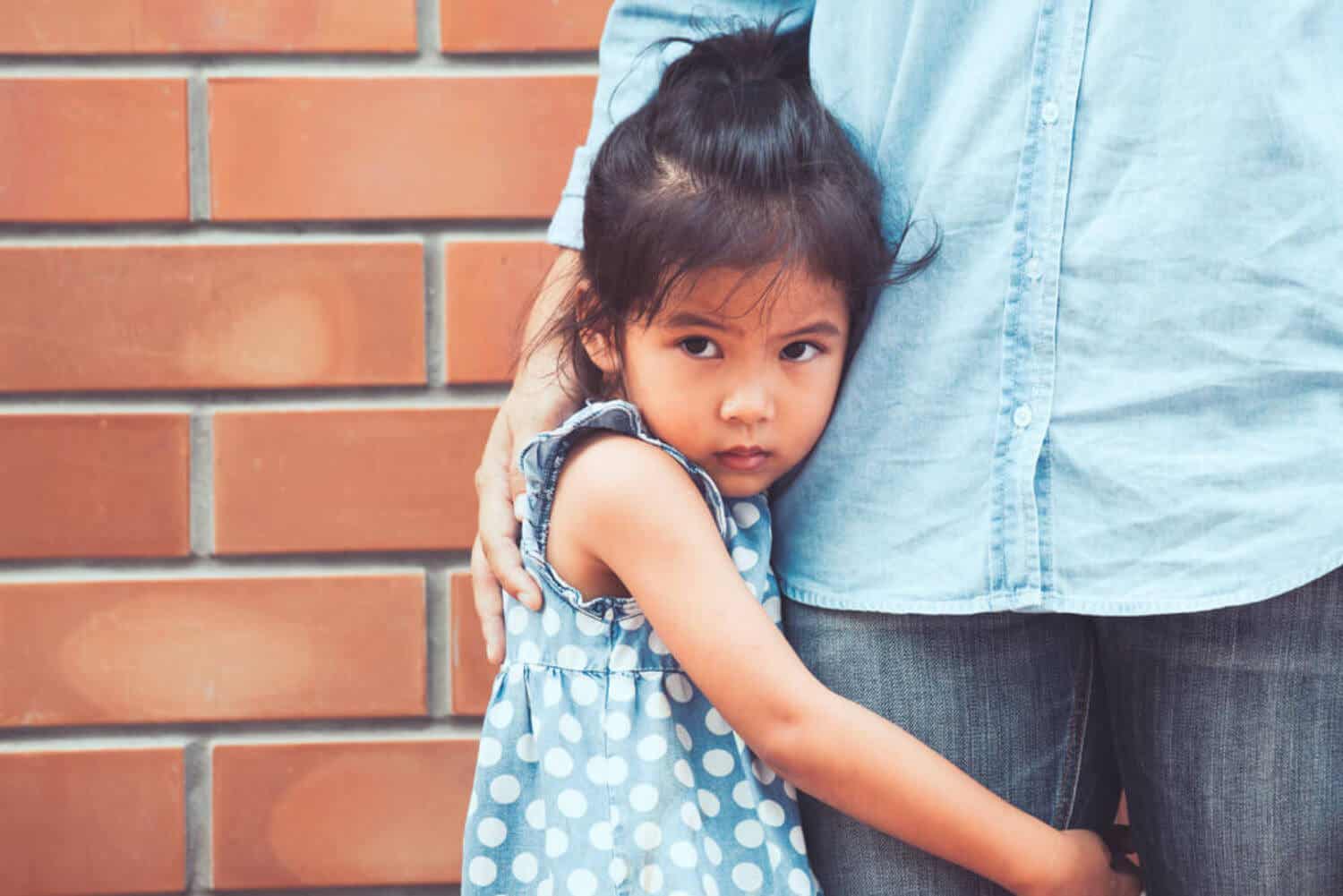 A small child hugging her parent's leg, looking afraid.