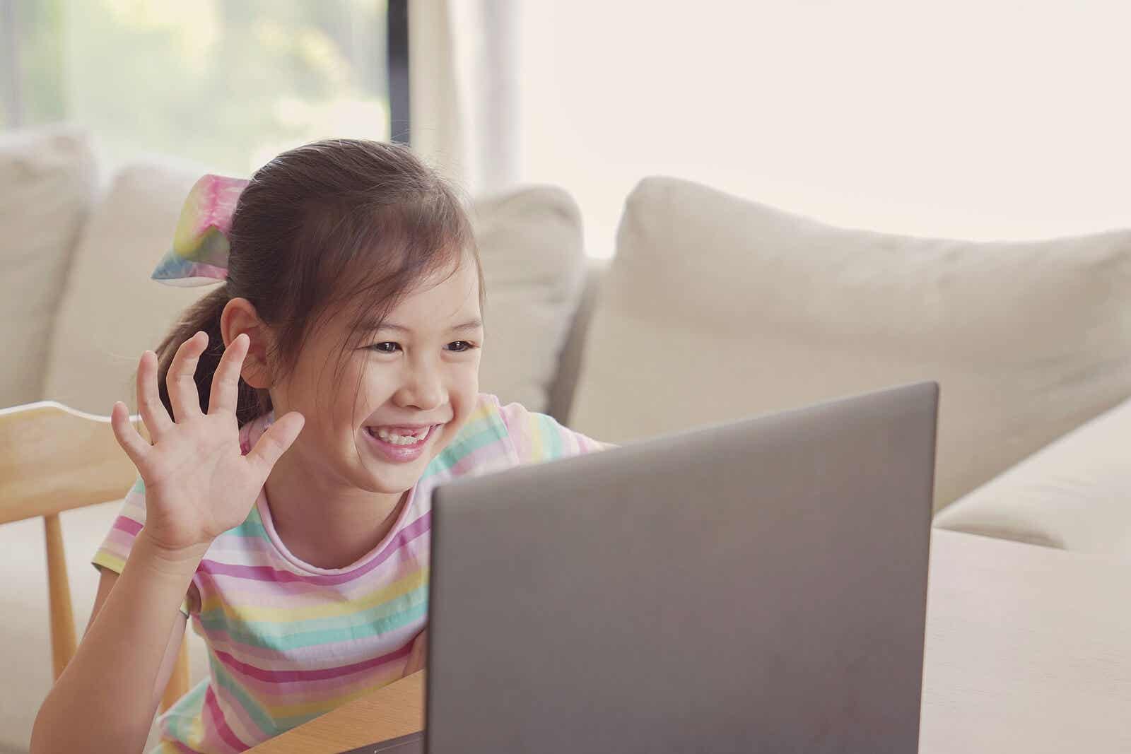 A young girl smiling and waving at the screen of her computer during online learning.