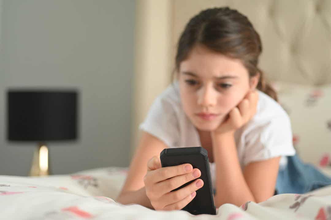 Een pre-tiener die op haar bed ligt en naar haar mobiele telefoon kijkt.