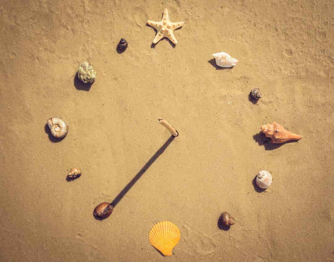 Een zelfgemaakte zonnewijzer met schelpen en een stok in het zand.