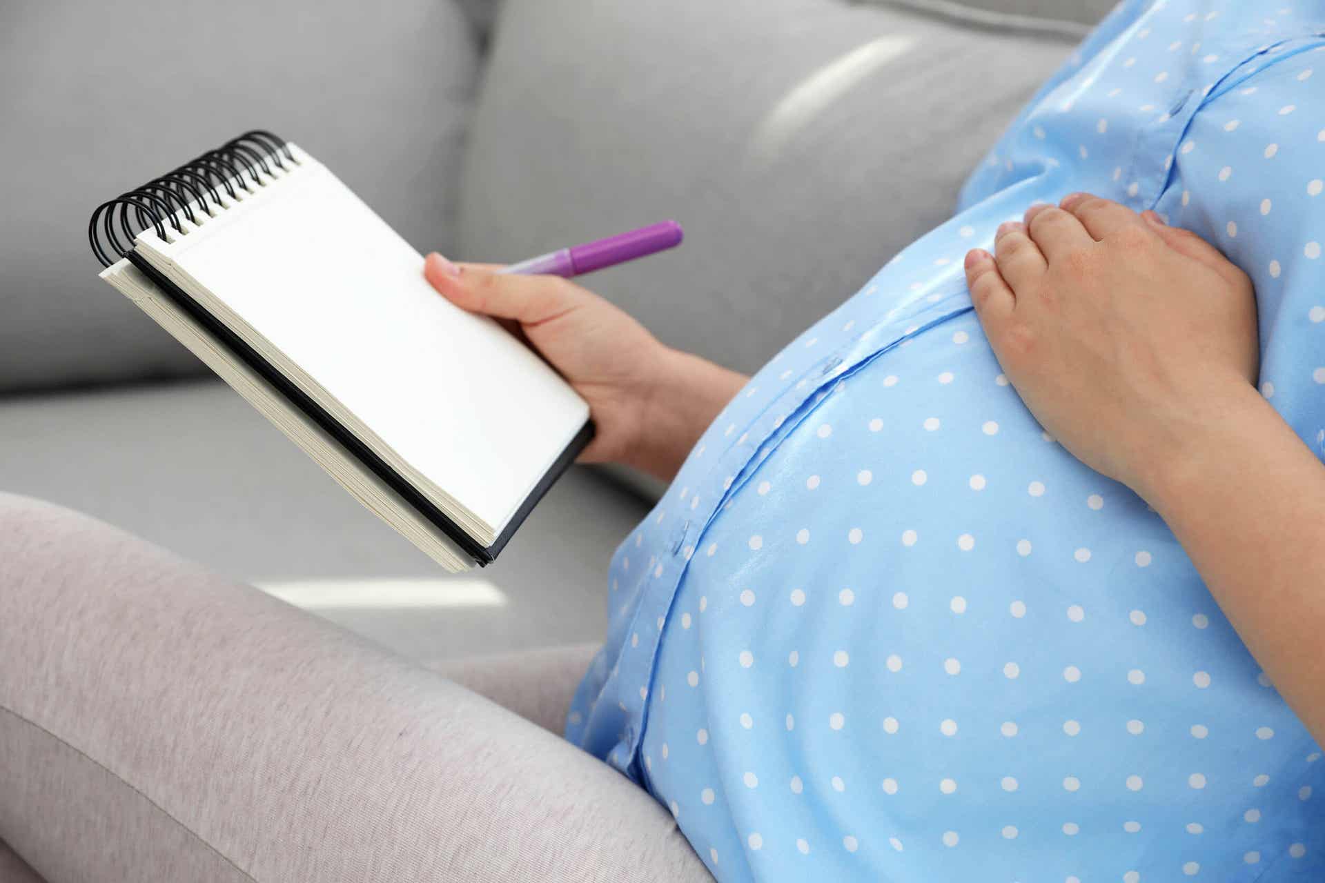 A pregnant woman toughing her belly with one hand, and holding a pen and notebook in the other hand.