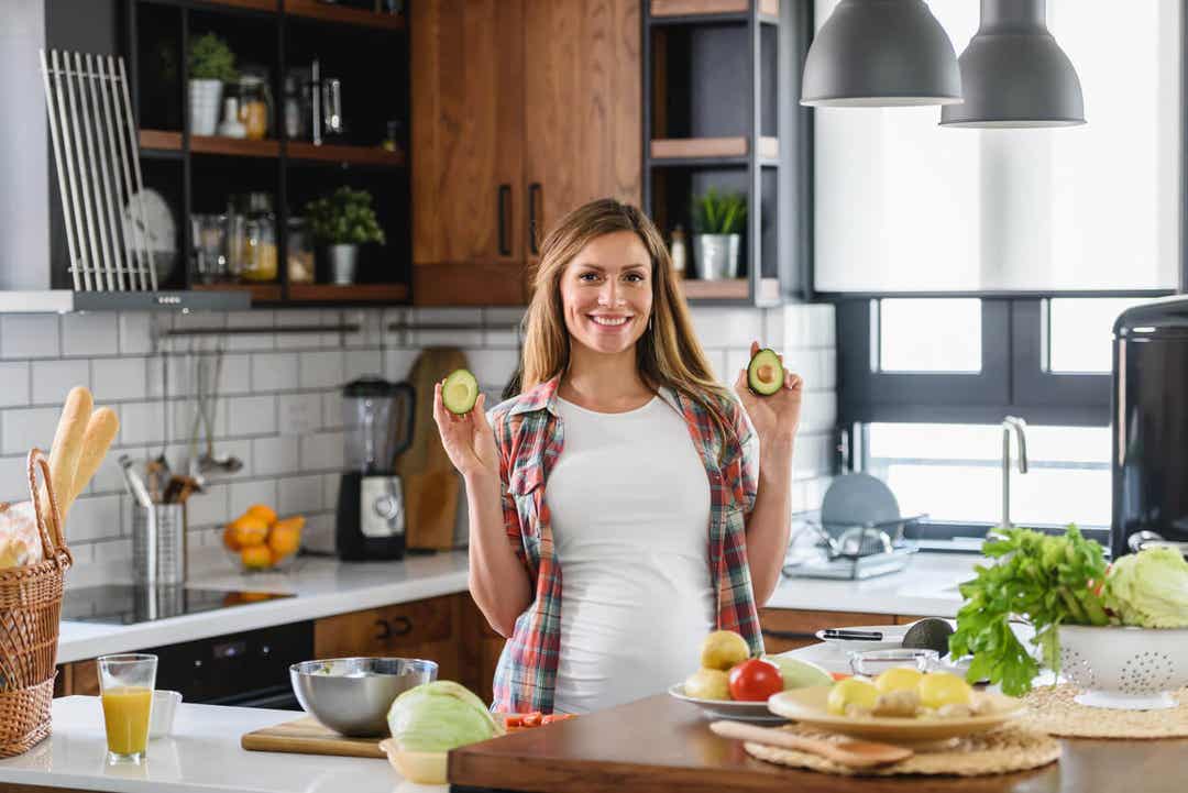 A pregnant woman in a kitchen holding two halves of an avocado and smiling.