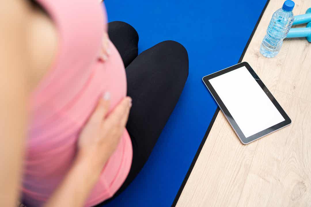 A pregnant woman exercising and looking at her tablet.
