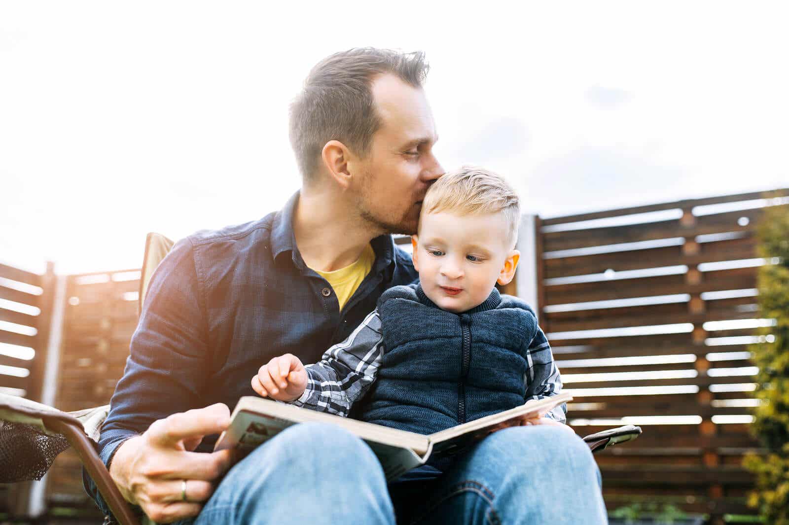 A father reading a book outdoors with his toddler son on his lap.