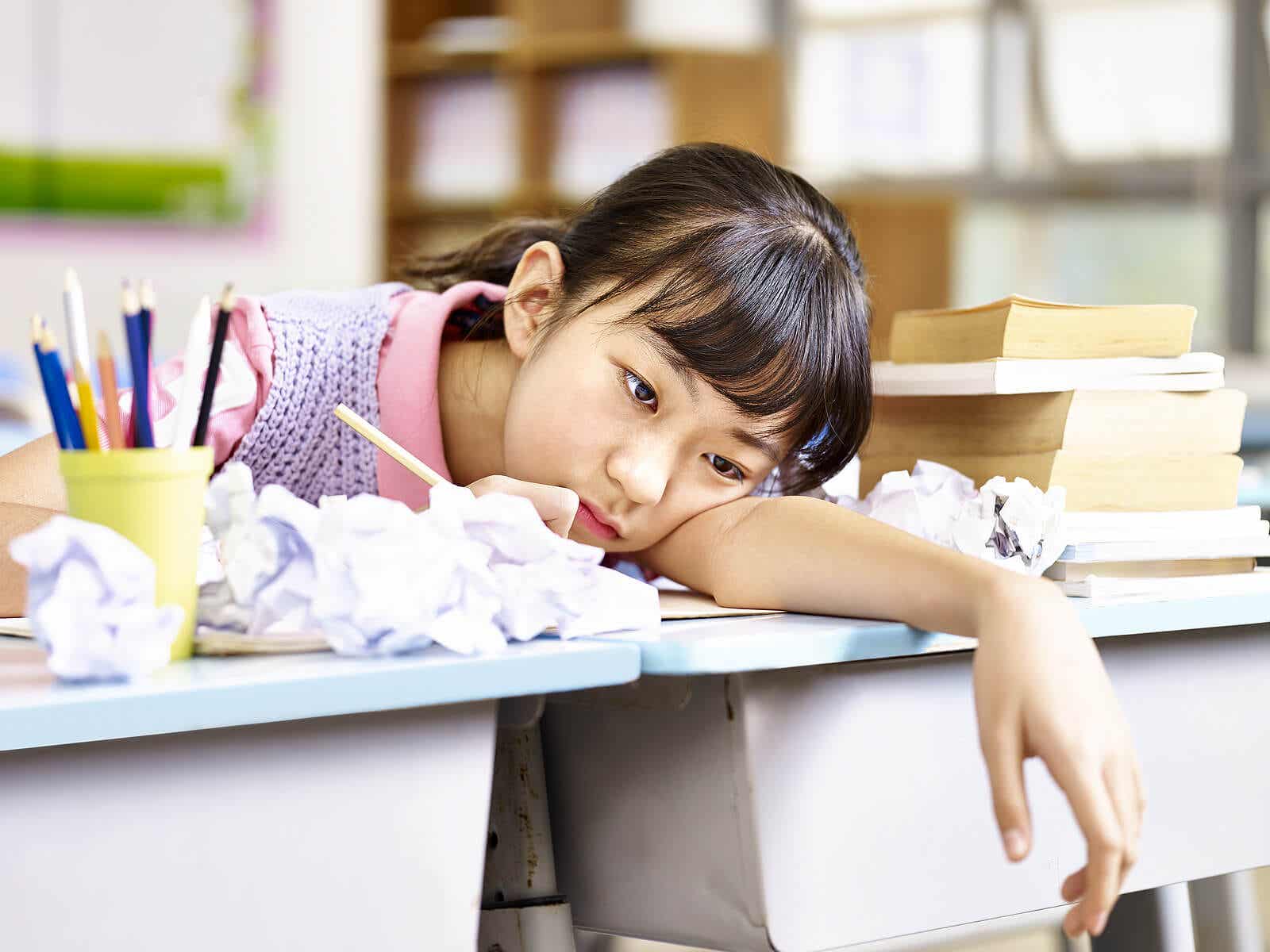 A preteen girl with her head on her school desk surrounded by crumpled up papers, looking bored.