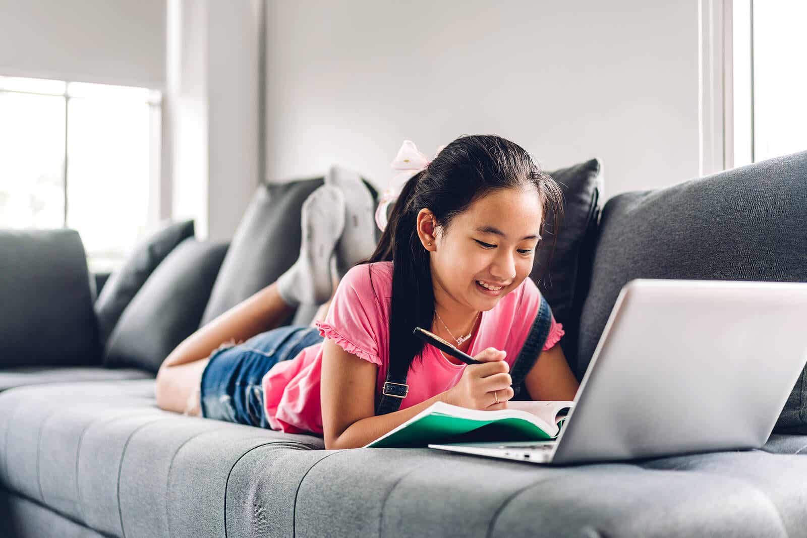 A child lying on a couch doing homework on her laptop computer.