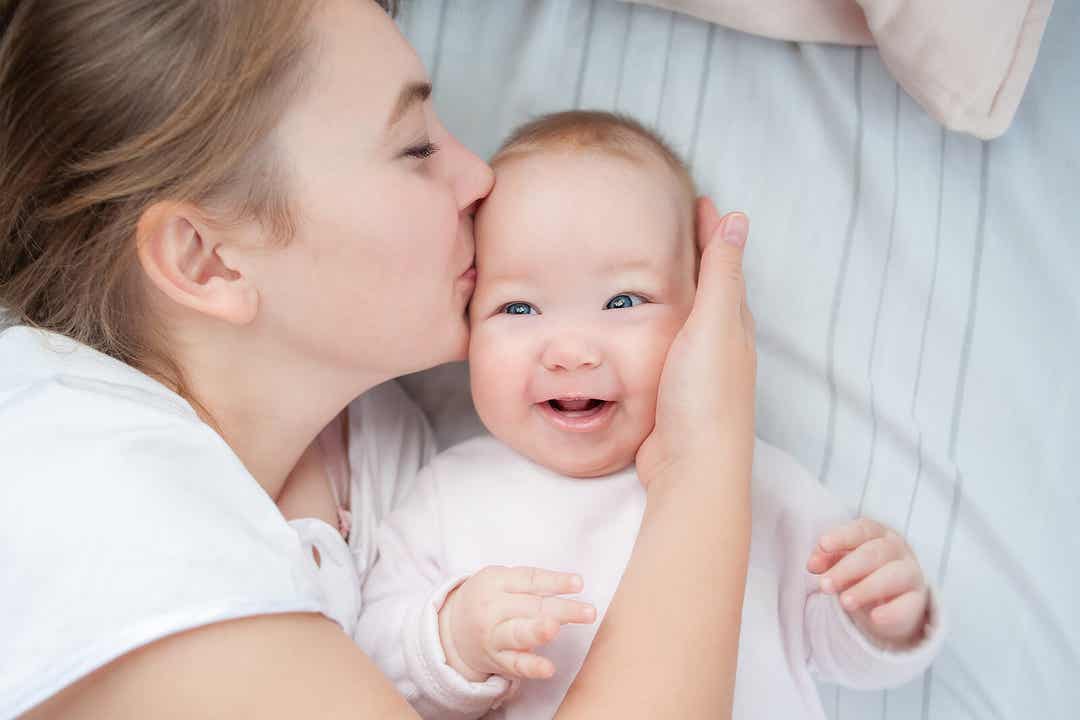 A mother lying in bed with her baby, giving her a kiss on the side of her head.