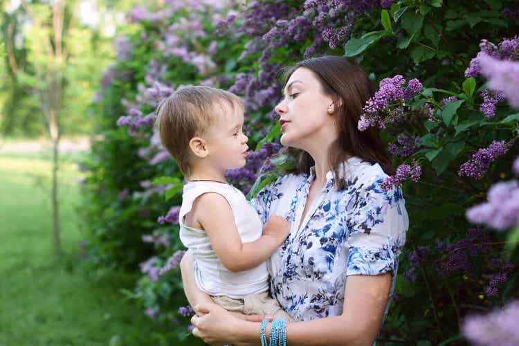 A woman holding her son in her arms near a lilac bush as they sing together.