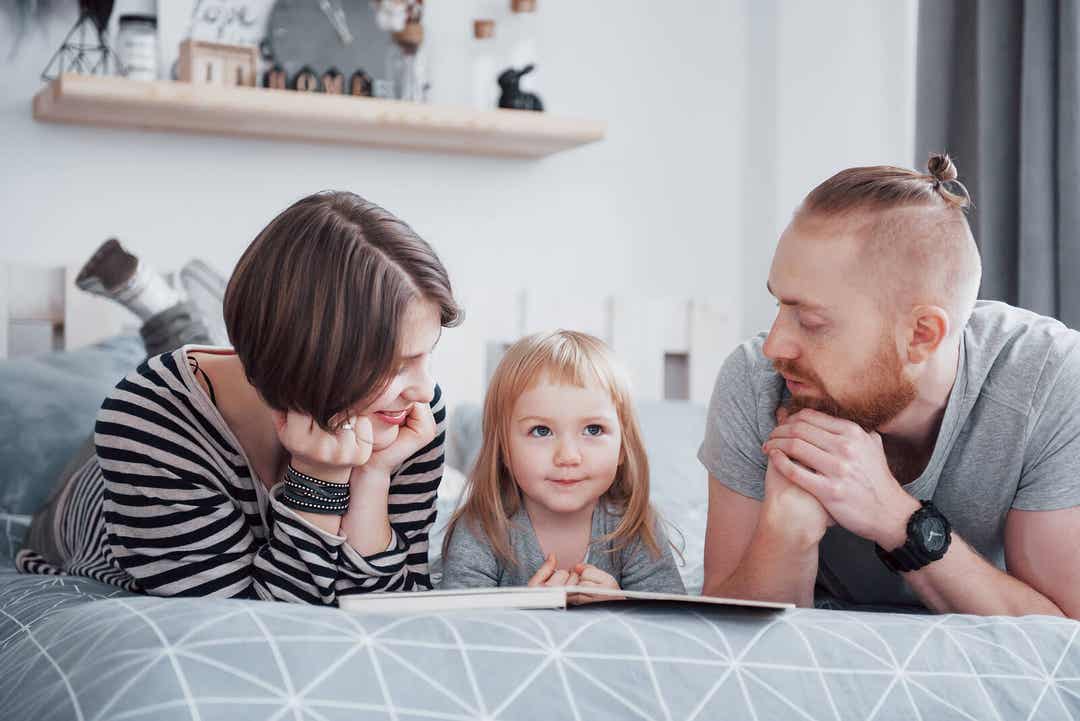 A mother and father lying on the couch reading a book with their toddler daughter.