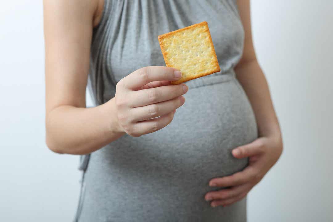 Een vrouw met een cracker in de ene hand en haar zwangere buik in de andere.