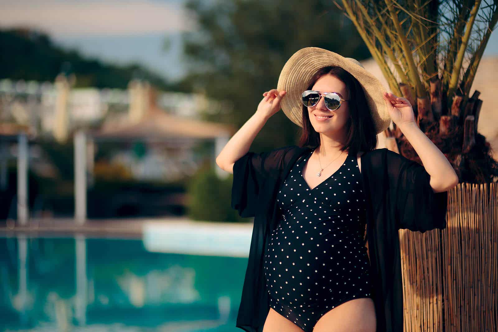 A pregnant woman standing in the sun by a pool.