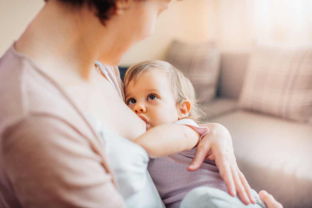 Een babymeisje dat borstvoeding geeft terwijl ze haar moeder in de ogen kijkt en haar hand op de andere borst van haar moeder legt.