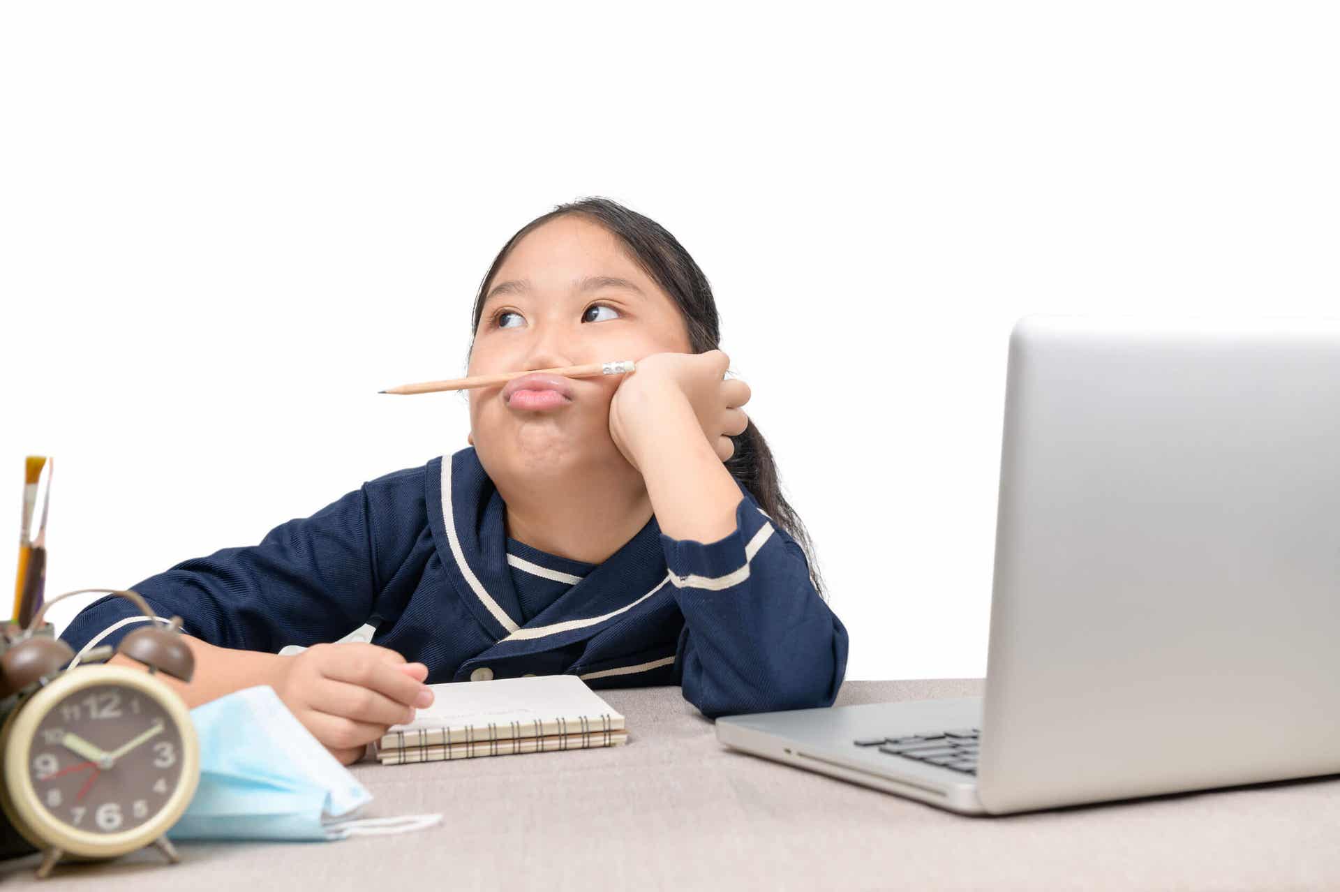 A girl sitting at a laptop computer, looking way and playing with a pencil.