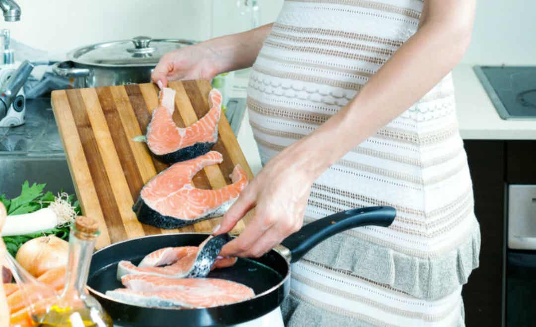 A pregnant woman cooking salmon in a skillet.
