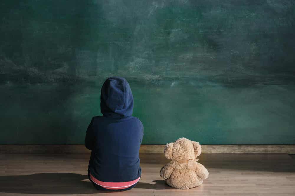 A child sitting on the floor next to his stuffed bear, staring at the wall.