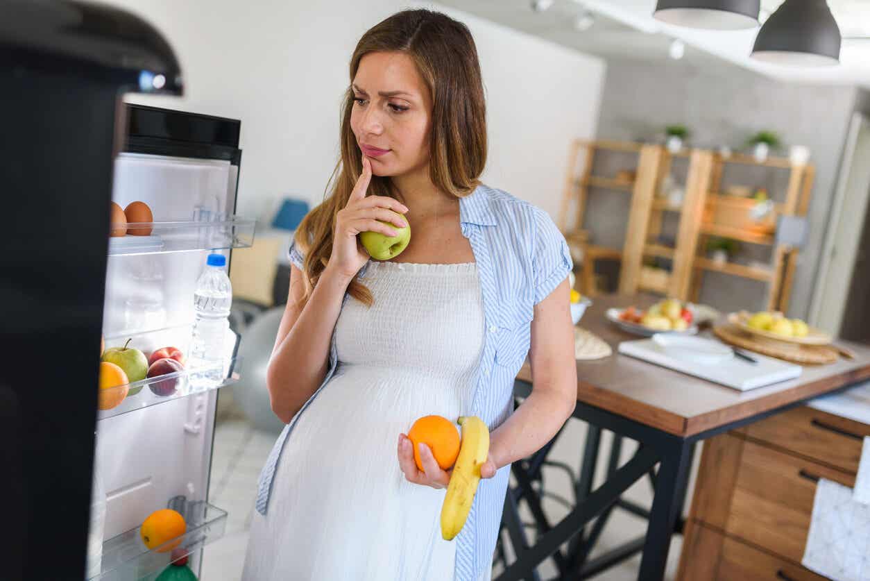 A pregnant woman grabbing fruit from the fridge.