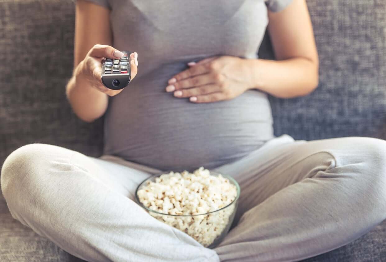 A pregnant woman eating popcorn and watching TV.