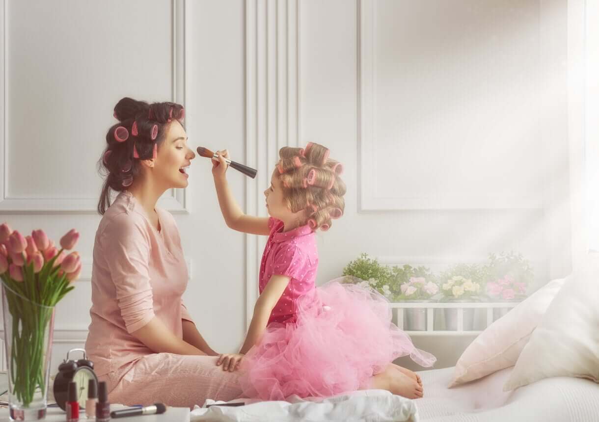 A daughter putting makeup on her mother's face while both of them wear curlers in their hair.