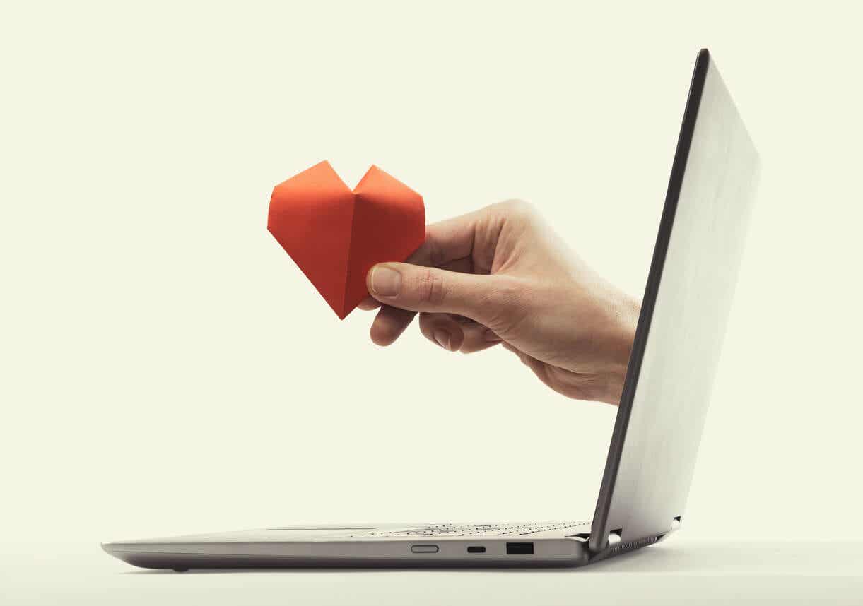 A hand reaching out from the screen of a laptop, holding a paper heart.