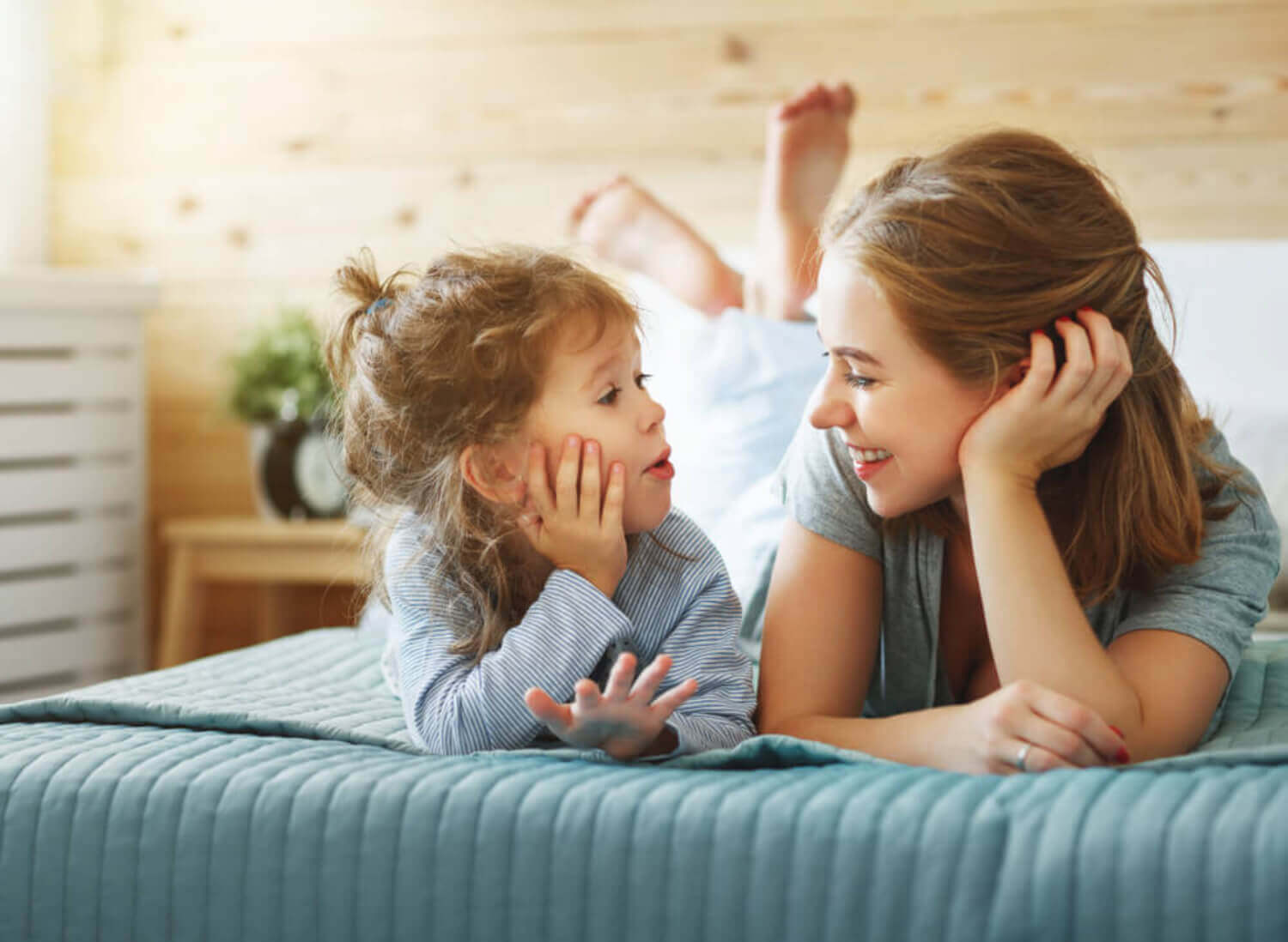 A mother and her toddler daughter lying in bed talking.