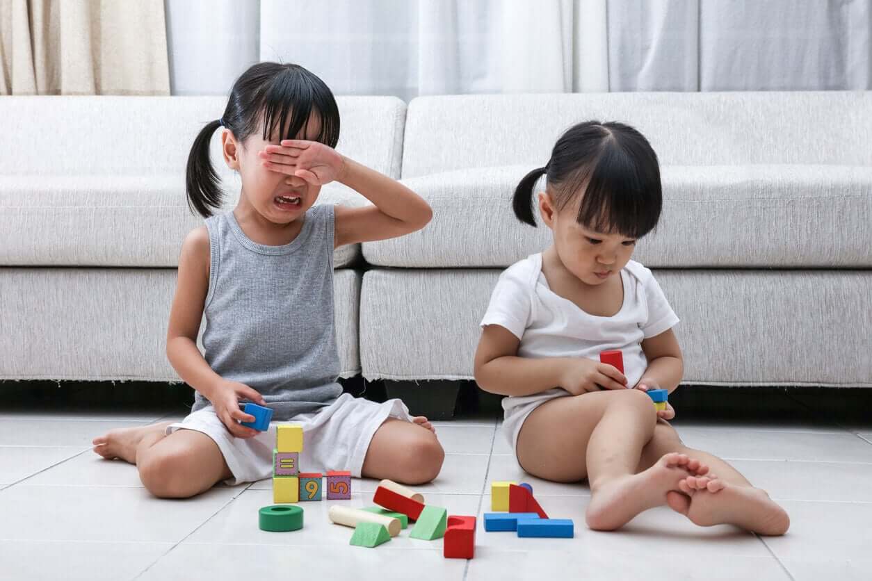 Two twins playing with blocks playing on the floor, one crying.