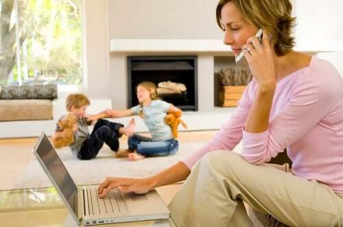 En kvinna pratar i telefon och arbetar på sin bärbara dator medan hennes barn stojar i bakgrunden.