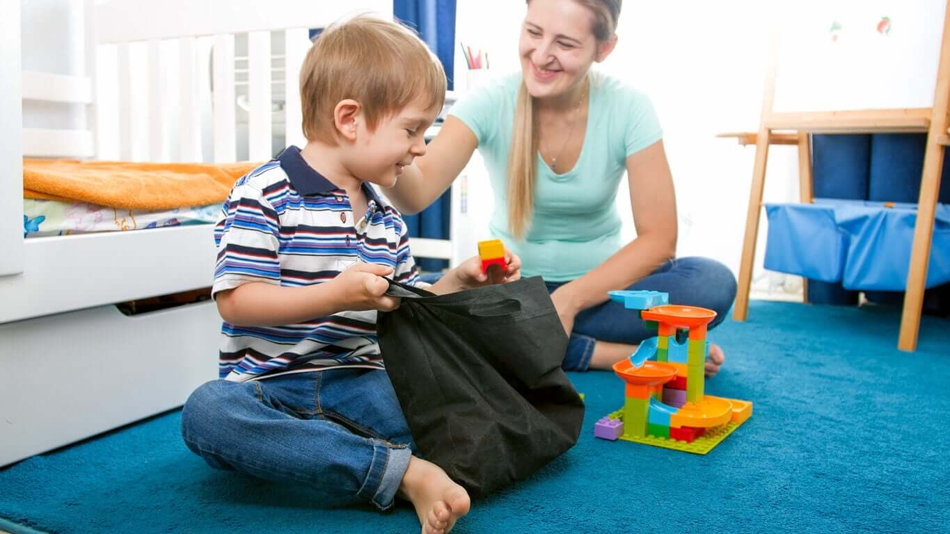 A child putting away toys in his room while his mother pats his back.