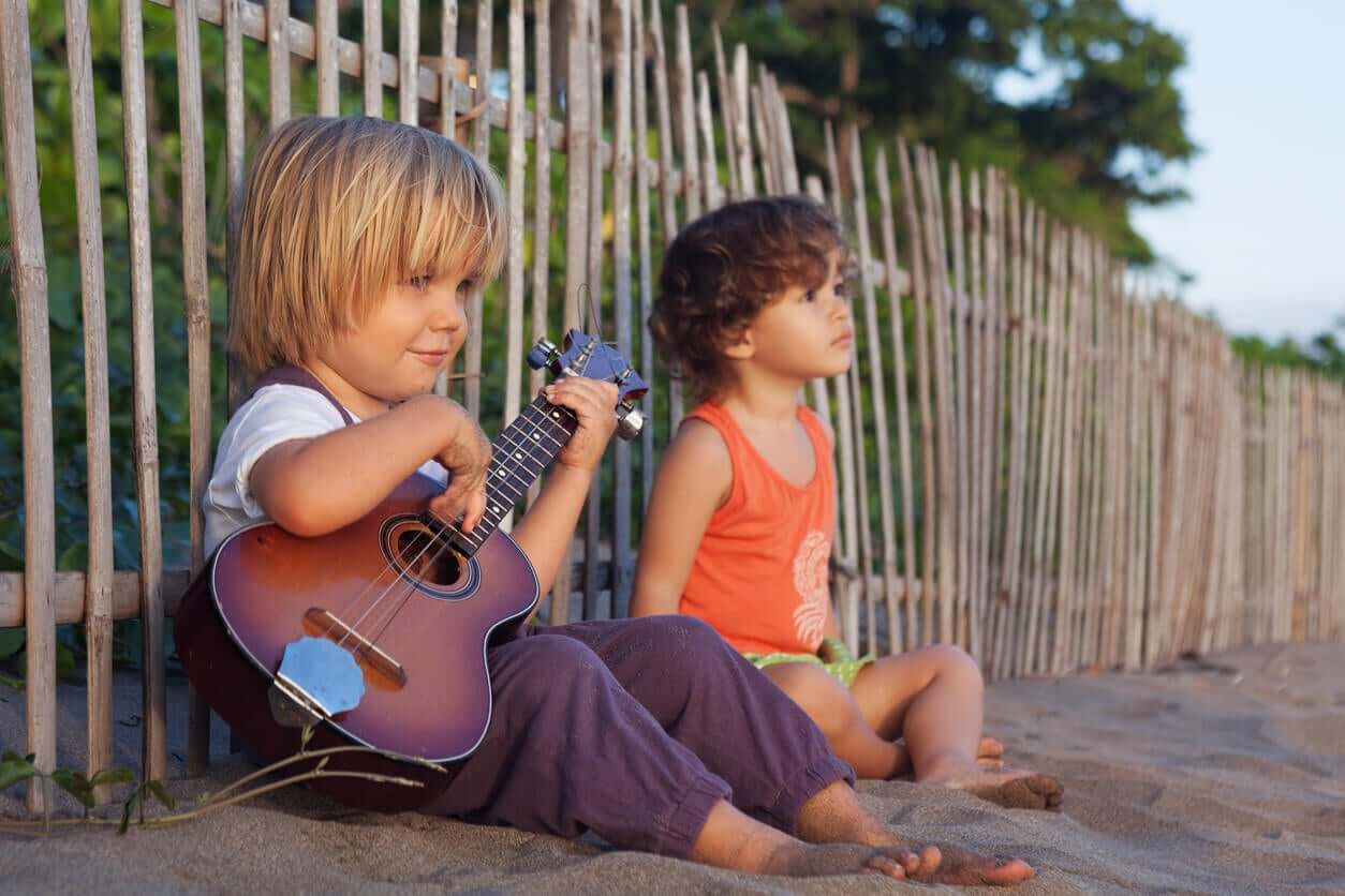 Two young boys sitting on the beach, one playing the ukelele.