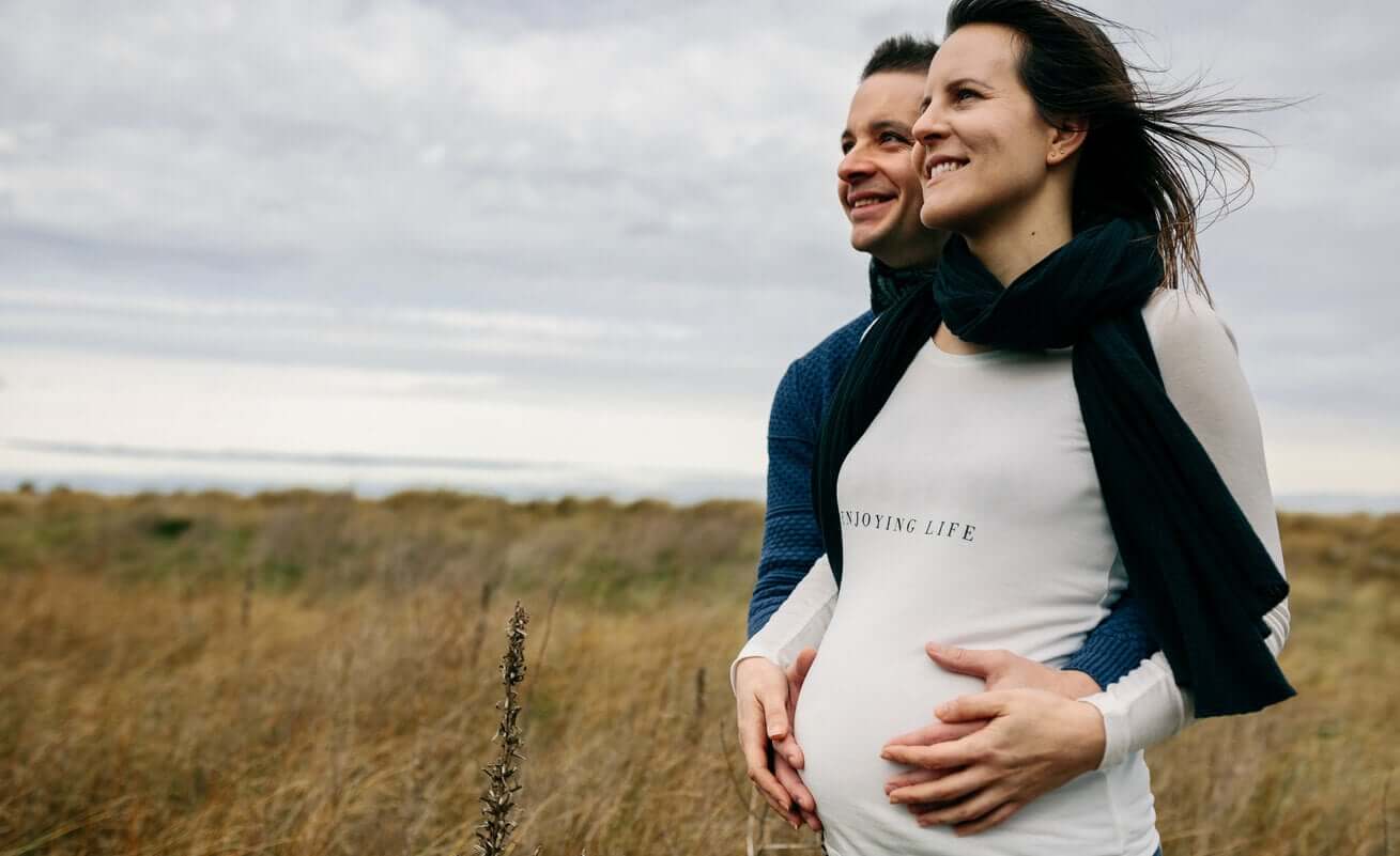 A pregnant couple standing in a field smiling.