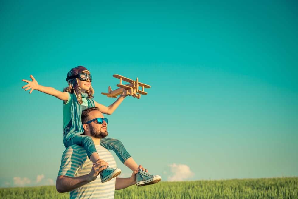 A girl dressed as an aviator and holding a wooden airplane while sitting on her father's shoulders.