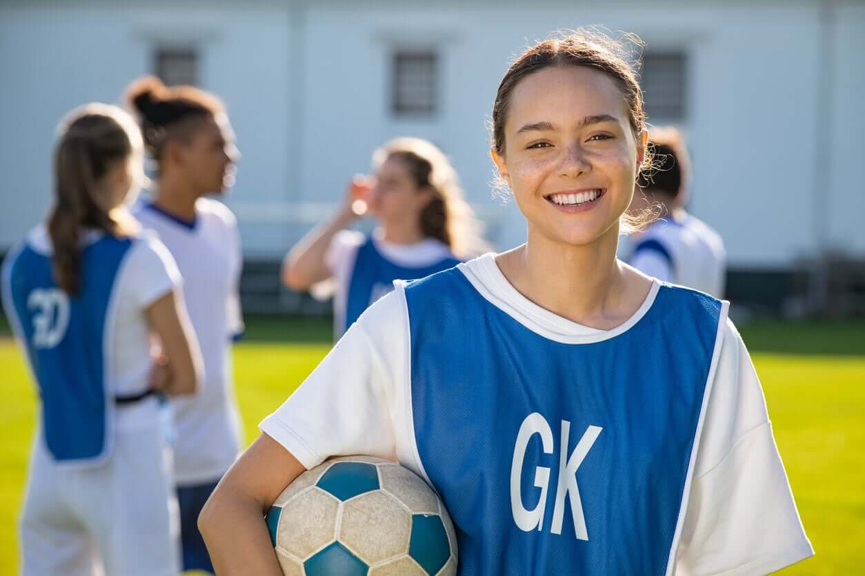 En tjej som bär en fotbollsuniform och håller en fotboll medan hon ler.