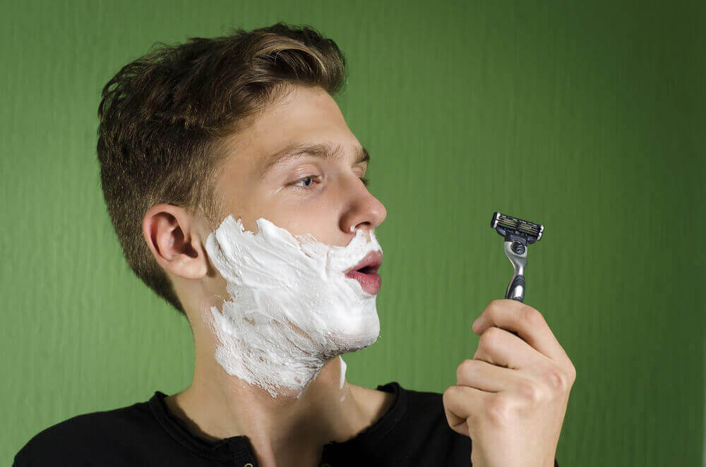 A teenager with shaving cream on his face looking at a razor.