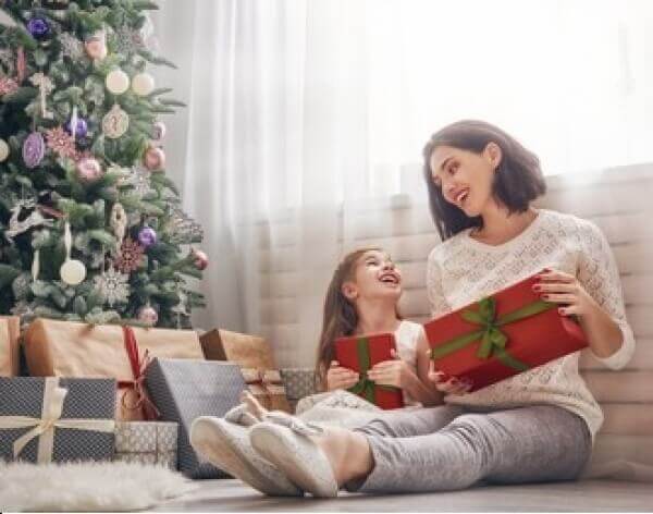 A mother and her daughter sitting on the floor by a Christmas tree and holding Christmas presents.