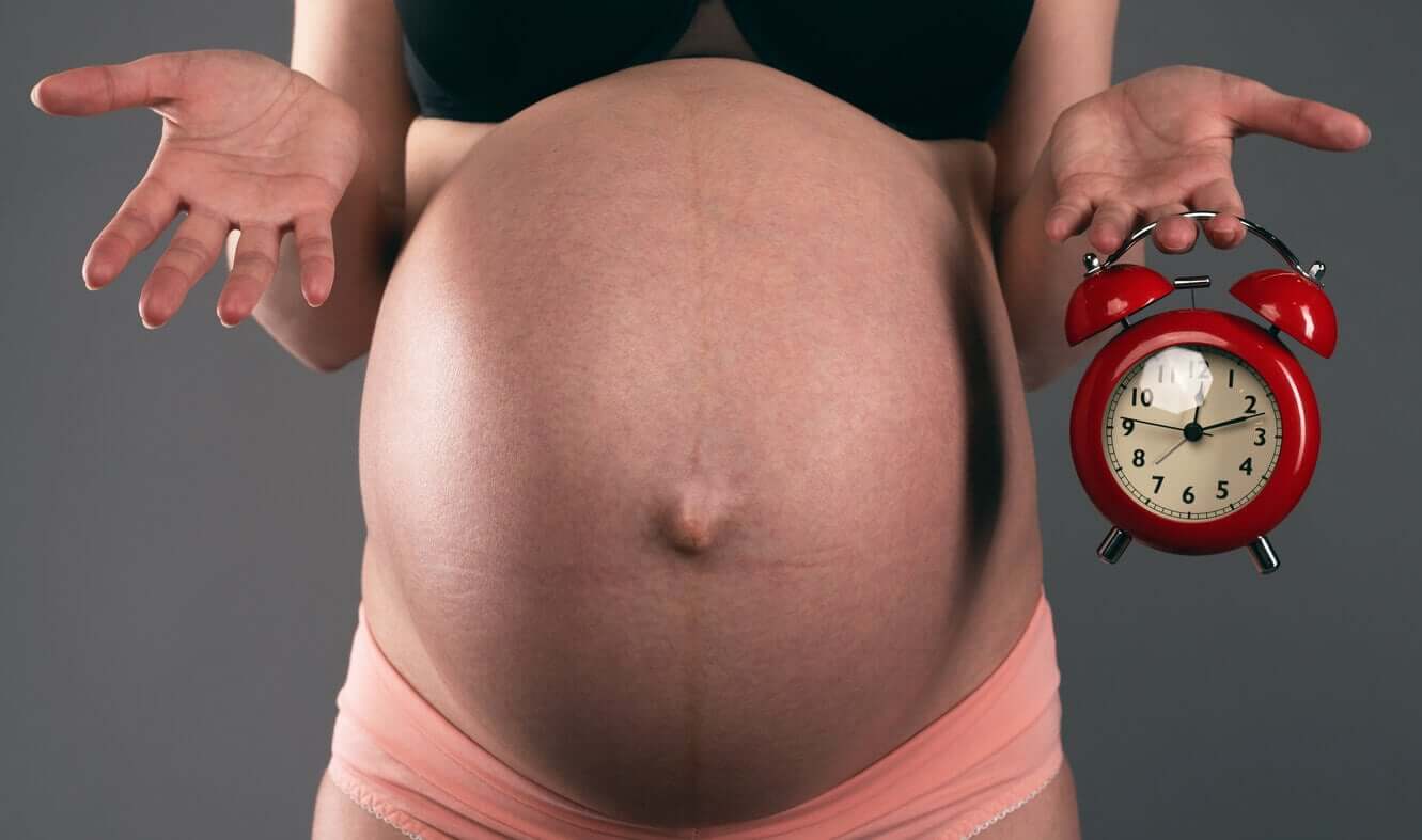A very pregnant woman holding an alarm clock, looking impatient.