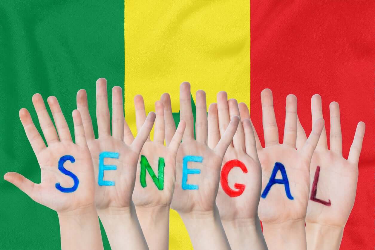 Hands with letters painted on them spelling the word Senegal, in front of the Senegalese flag.