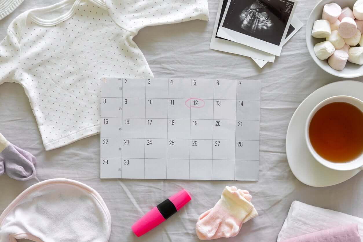 A calendar surrounded by a newborn baby tee, a bib, tiny socks, and ultrasound pictures.