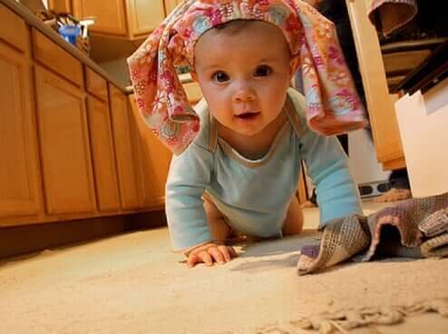 A baby crawling on the kitchen floor while wearing a pair of pants on her head like a hat.