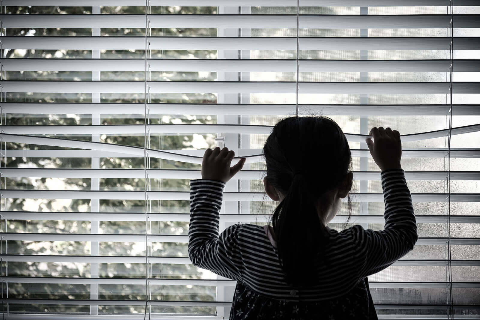 En liten flicka tittar ut genom persiennerna i ett fönster.