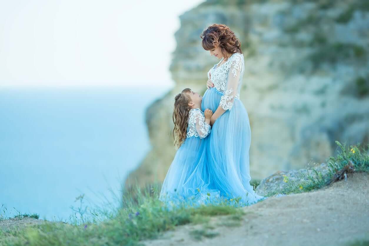 A pregnant woman and her little girl smiling at one another while standing on a cliff over the ocean.