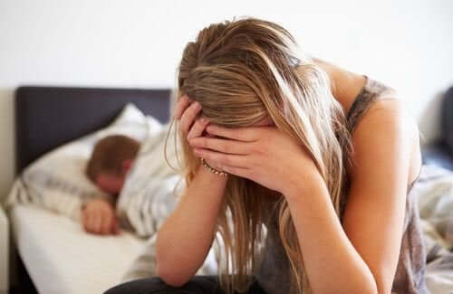 A woman sitting on the edge of her bed looking frustrated and tired while her partner sleeps.