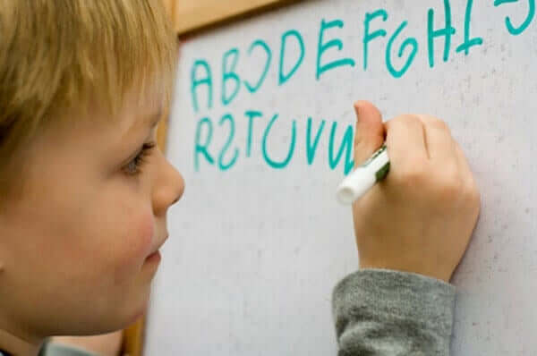 A small child writing the alphabet on a whiteboard, with many letters written backwards.