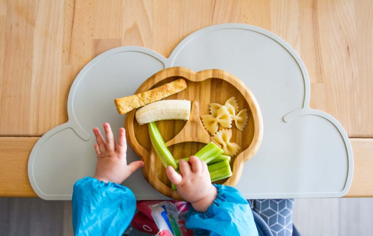 A baby eating from a plate with a banana, vegetable ssticks, bowtie pasta, and a breadstick.