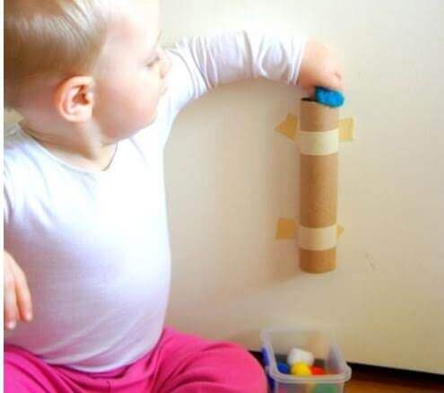 A baby pushing pom-poms through a cardboard tube tables to the wall.