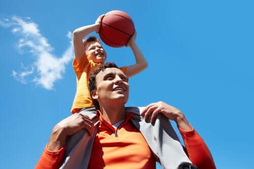 A child sitting on his father's shoulder's and throwing a basketball.