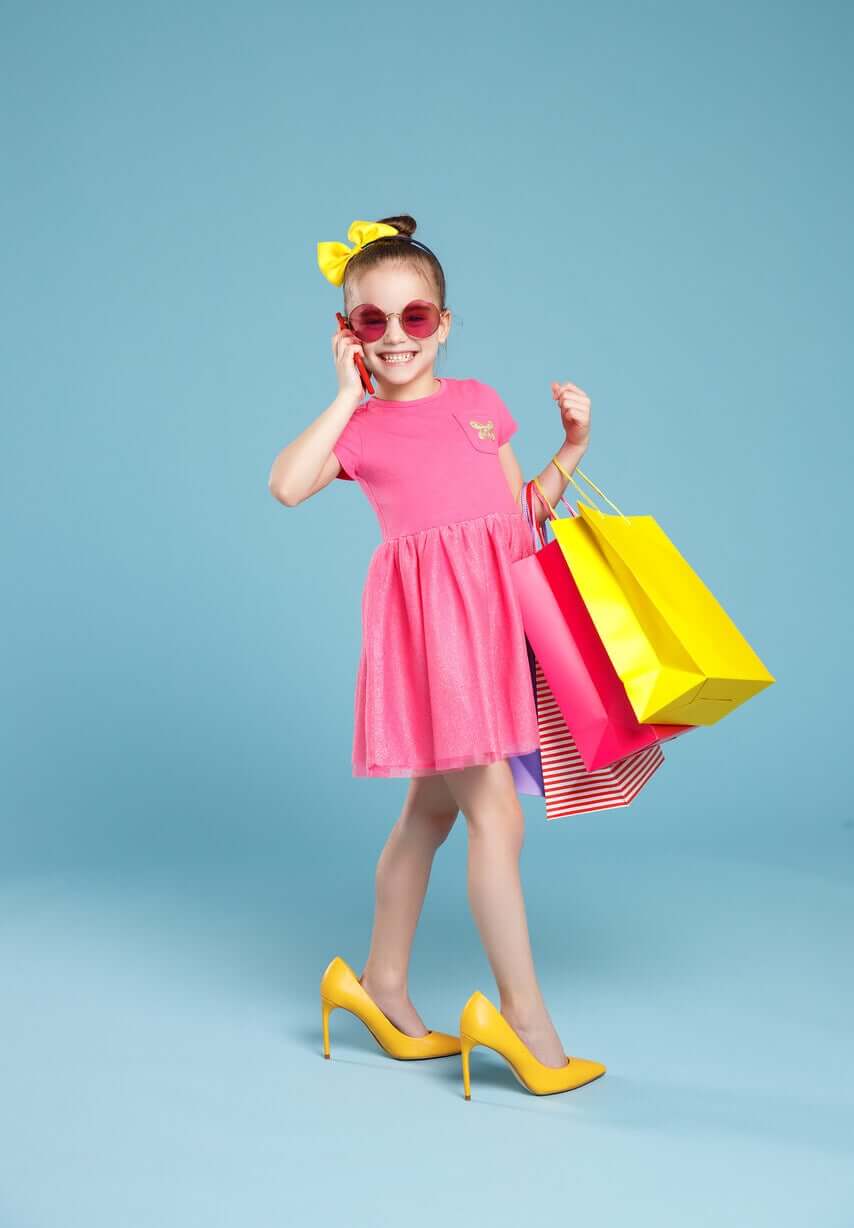 A young girl wearing a dress, sunglasses, and high heels while carrying shopping bags and using a cell phone.