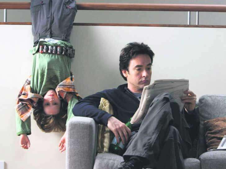 A child hanging upside down on a railing behind a man sitting on a couch.