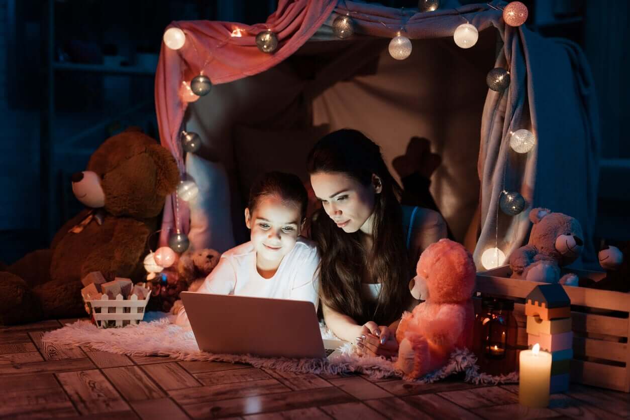 A mother and her daughter laying in a fort and watching a movie on a laptop screen.